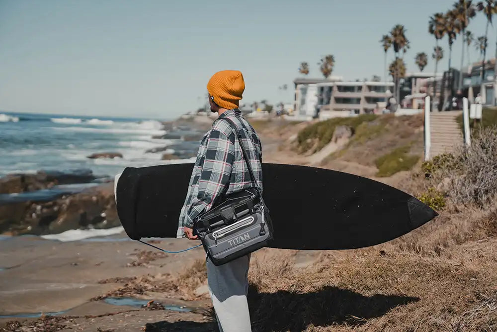 Guy at the beach with a surf board and Titan 40 Can Collapsible Cooler